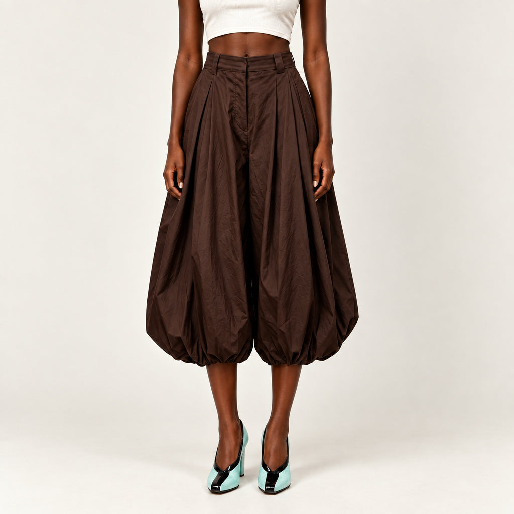 A model wearing high-waisted, voluminous brown pleated culotte-style pants with a cropped white top and mint green heels, shown from the waist down against a plain off-white studio background.