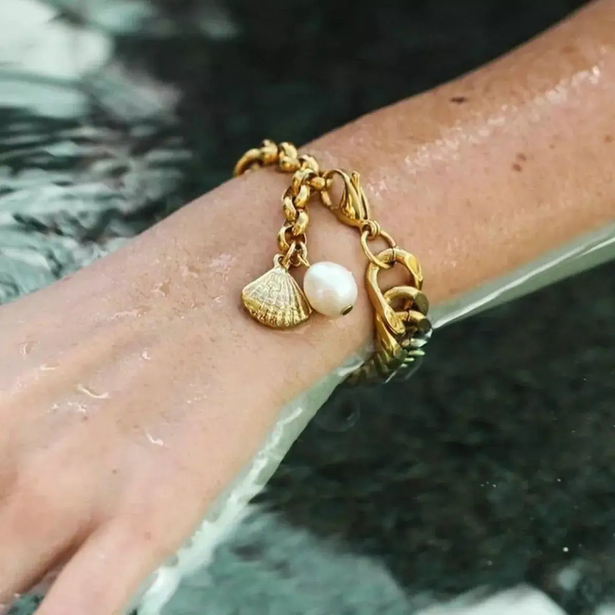 Gold bracelet with shell and pearl charm on a wrist by water