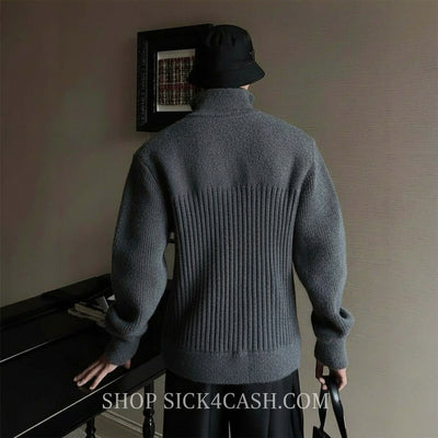 Person wearing a gray sweater and black hat, standing in a room with a piano and framed picture on the wall.