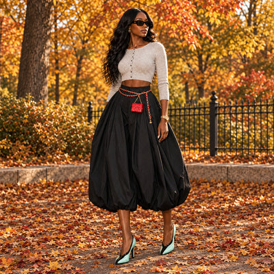 A person wearing a voluminous black high-waisted harem-style skirt with a gold chain belt and a small red crossbody bag, paired with mint green and black heels, standing on black diamond-plate steps in front of a cream paneled door.